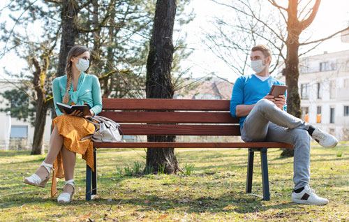 Social distancing: young man and woman, both with face coverings, sitting at opposite ends of a long park bench